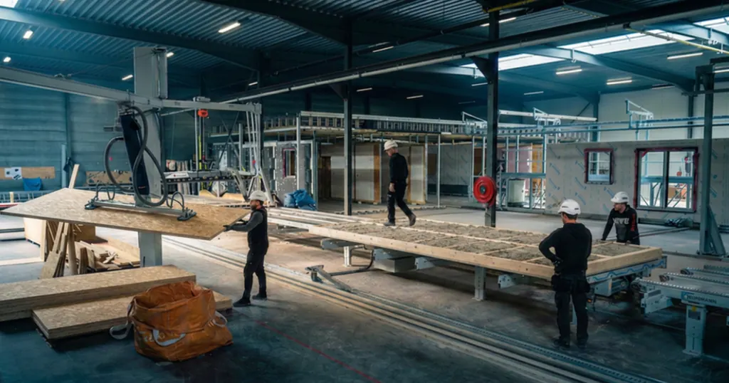 Inside the production workshop: hard-hatted workers handle a large frame panel with a vacuum lifter over an assembly table, stacked OSB boards in the foreground.